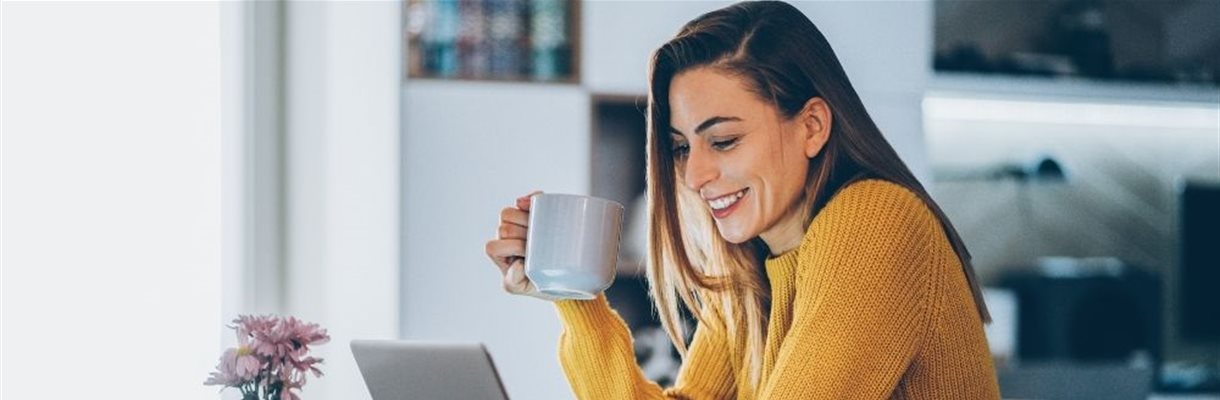 Dame is aan het thuiswerken met een laptop op de keukentafel en een kop koffie in haar hand